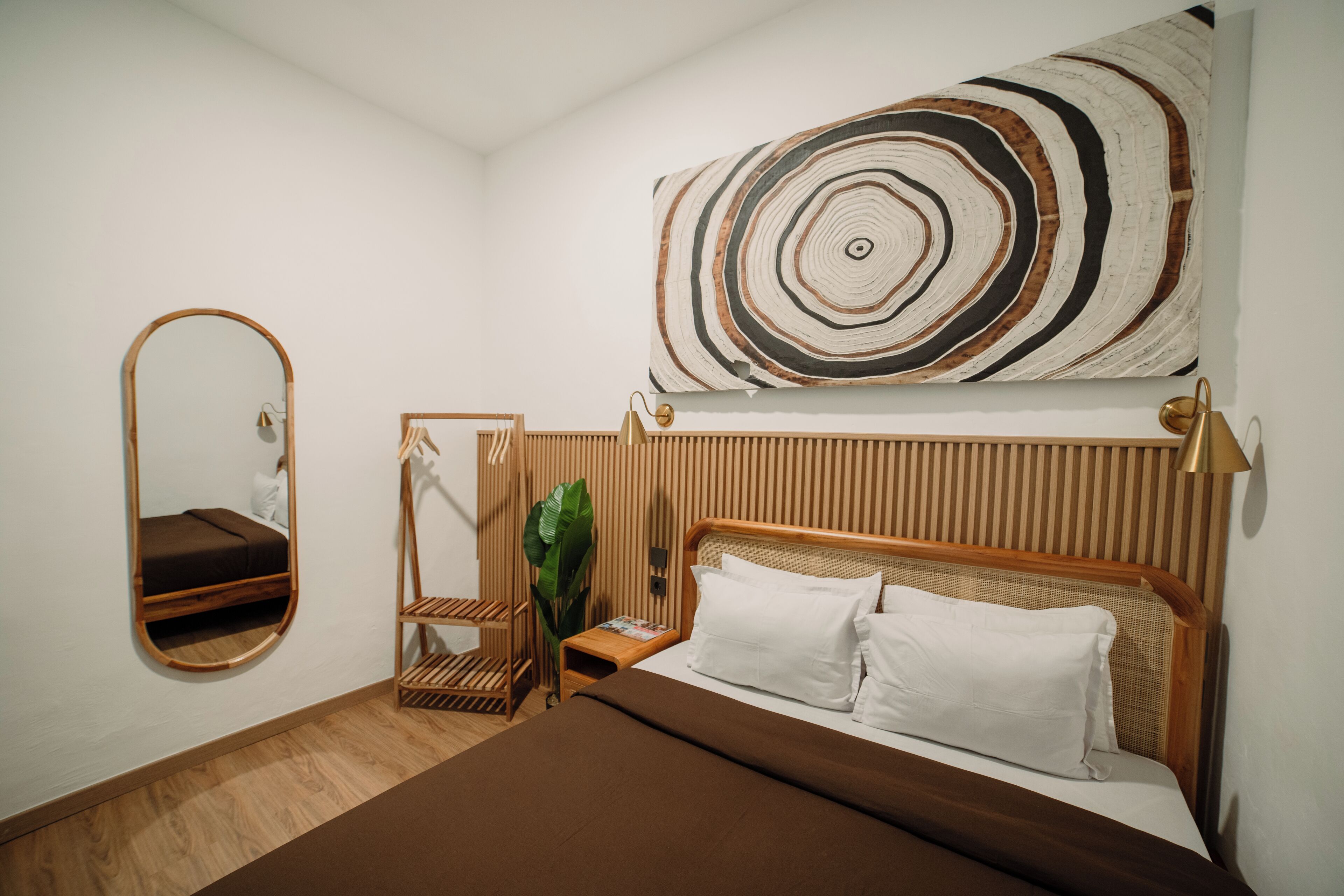 A bedroom with a bed, a bedside table, a mirror, a wall-mounted artwork, and a plant.