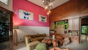 A living room with a beige sofa, a green bean bag, and a wooden coffee table. There are two panda paintings on the pink wall. - Snooze Hostel Malang (Malang)