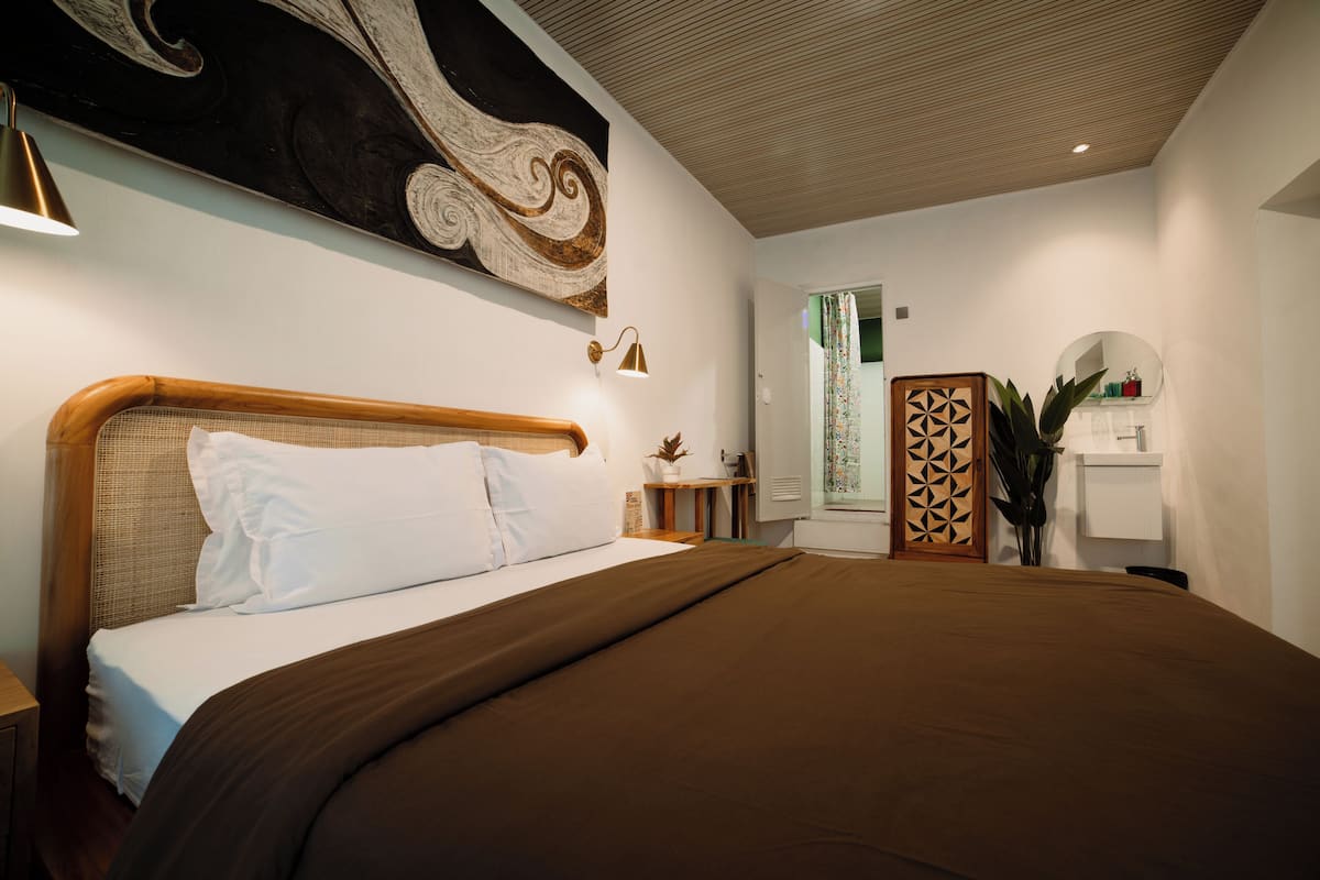 A bedroom with a large bed, a headboard, a bedside table, a wall-mounted lamp, a decorative wall piece, and a door with a geometric pattern.