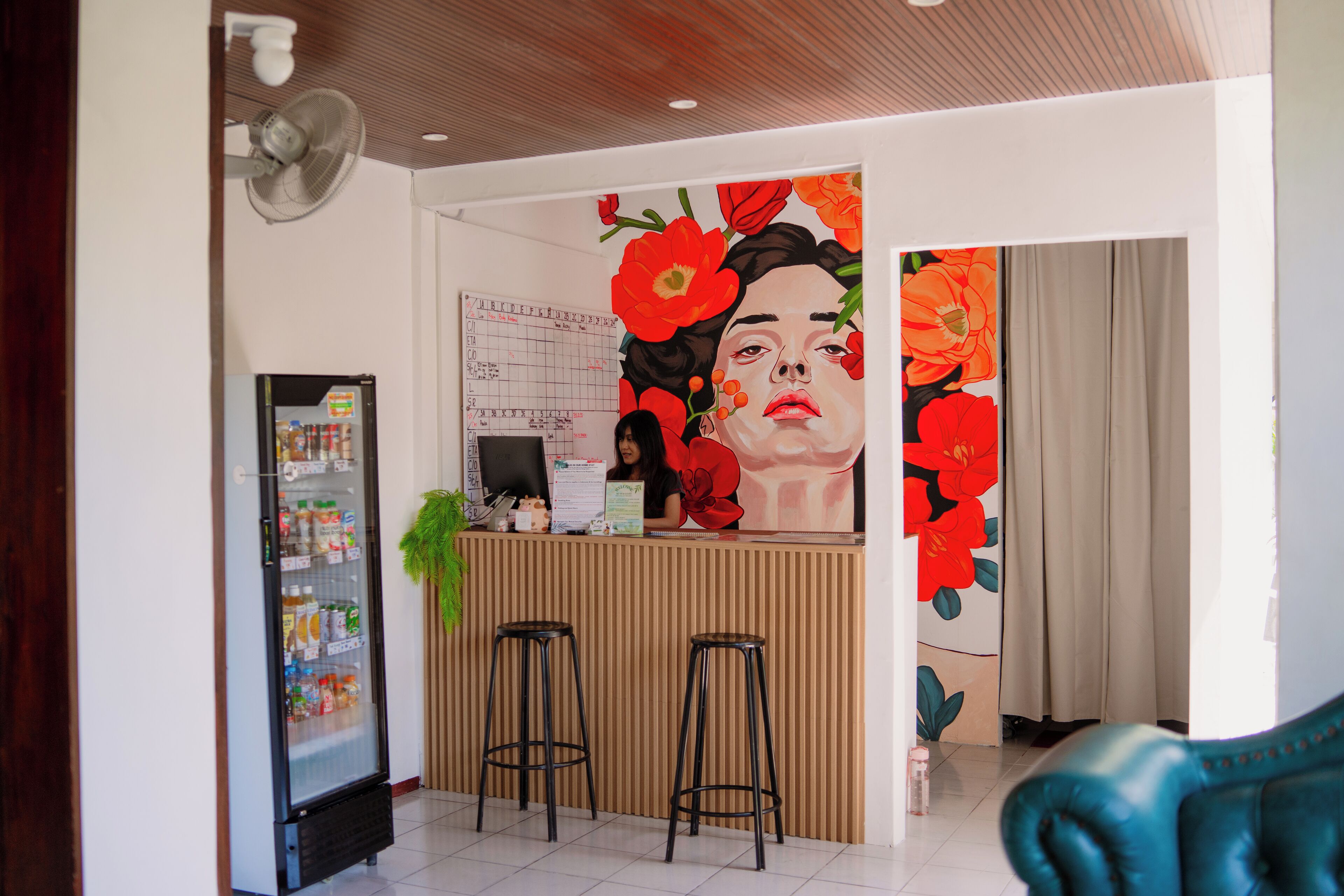 Un espace d’accueil avec un comptoir en bois, deux tabourets de bar, un réfrigérateur rempli de boissons et une grande fresque florale sur le mur.