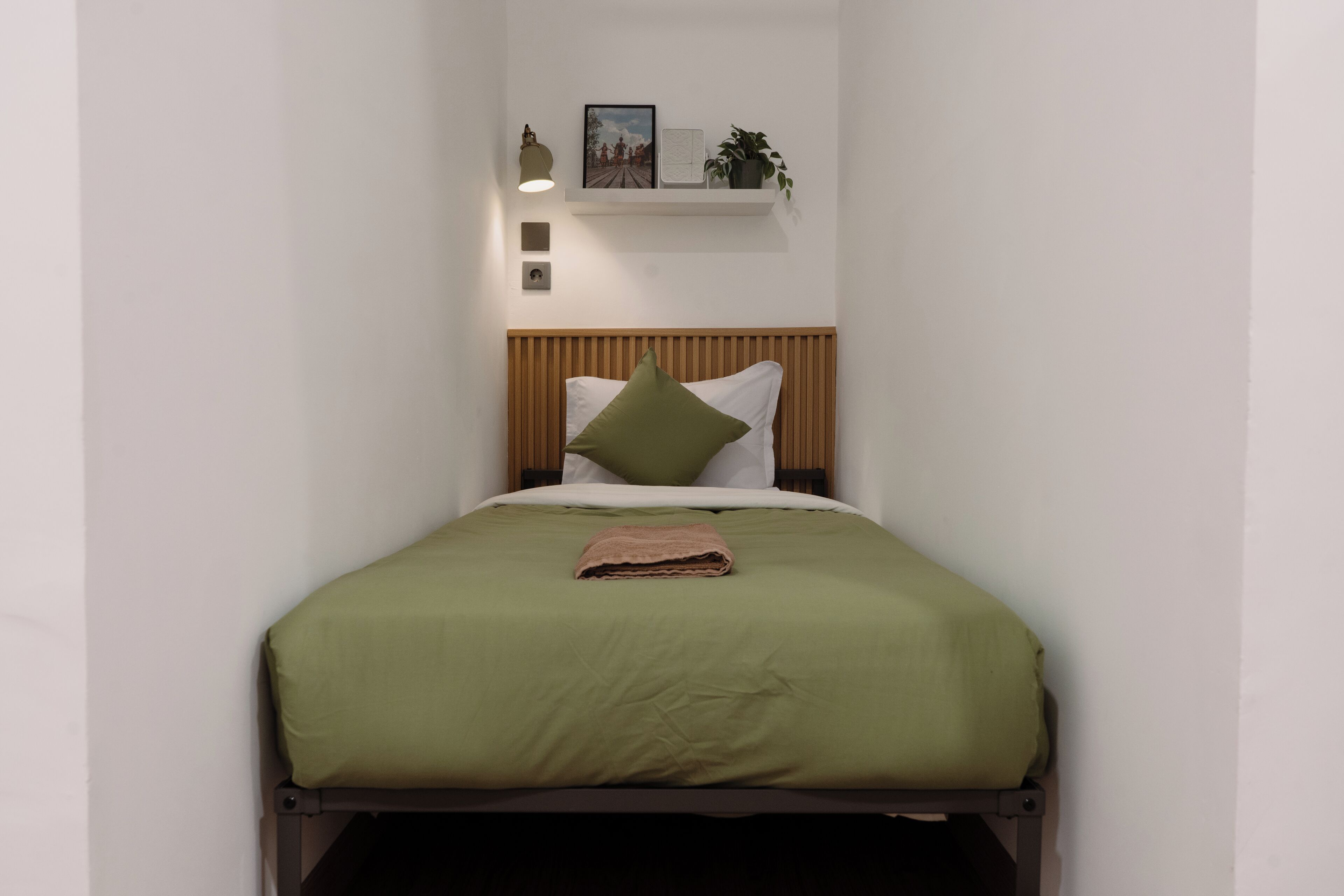 A neatly made bed with a green bedspread and a folded brown towel, a wooden headboard, and a shelf above the bed with a framed picture, a small plant, and a lamp.