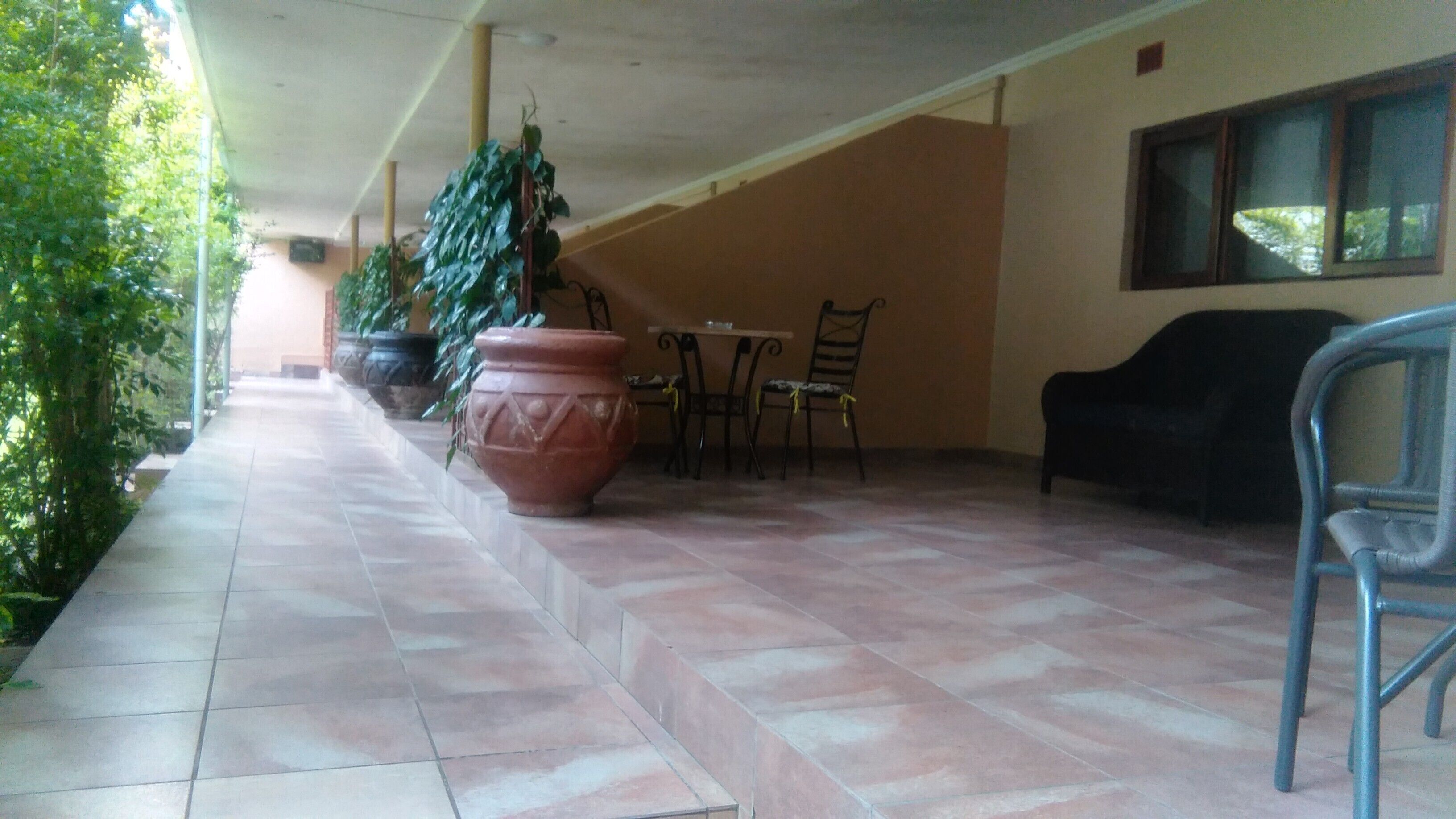 Un área de patio techada con suelo de baldosas, una mesa con sillas, una maceta de terracota grande y plantas en macetas.