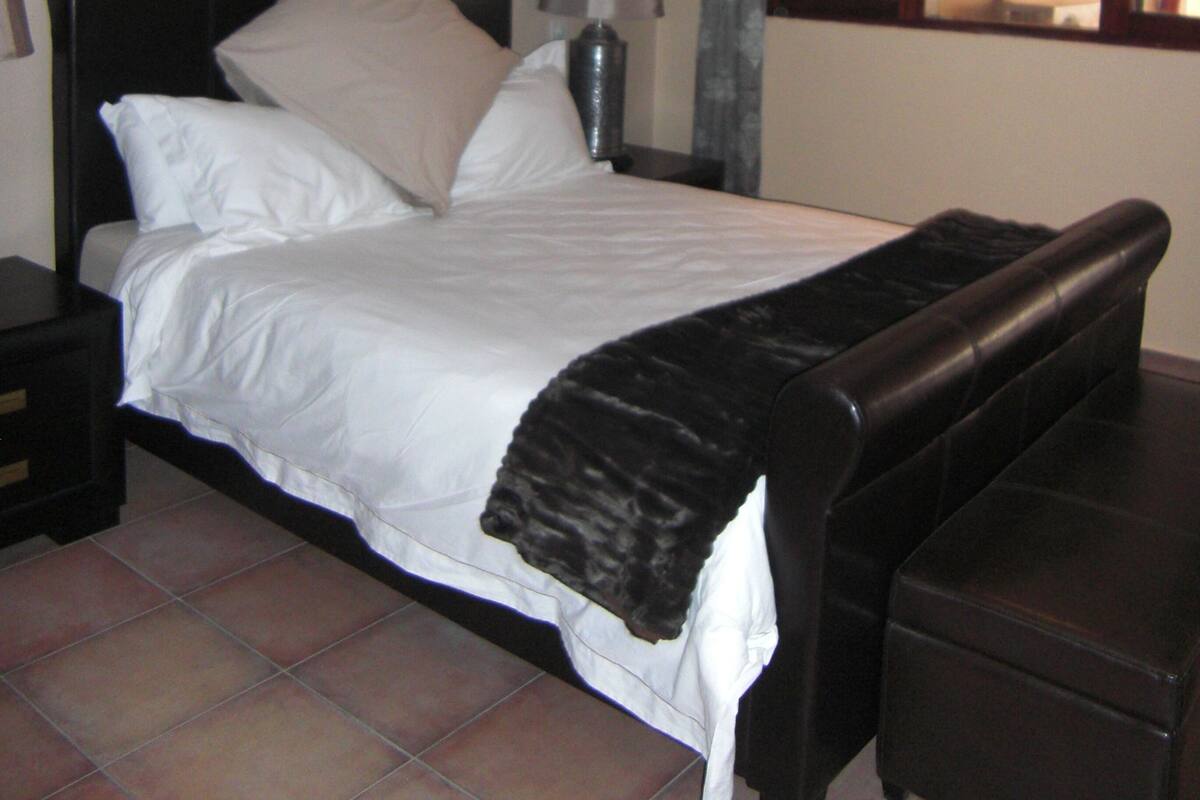 Un dormitorio con una cama, una mesita de noche, una lámpara, un banco y un espejo.