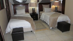 A hotel room with two beds, a nightstand, a lamp, and a small ottoman. - Shamba Lodge (Kitwe)
