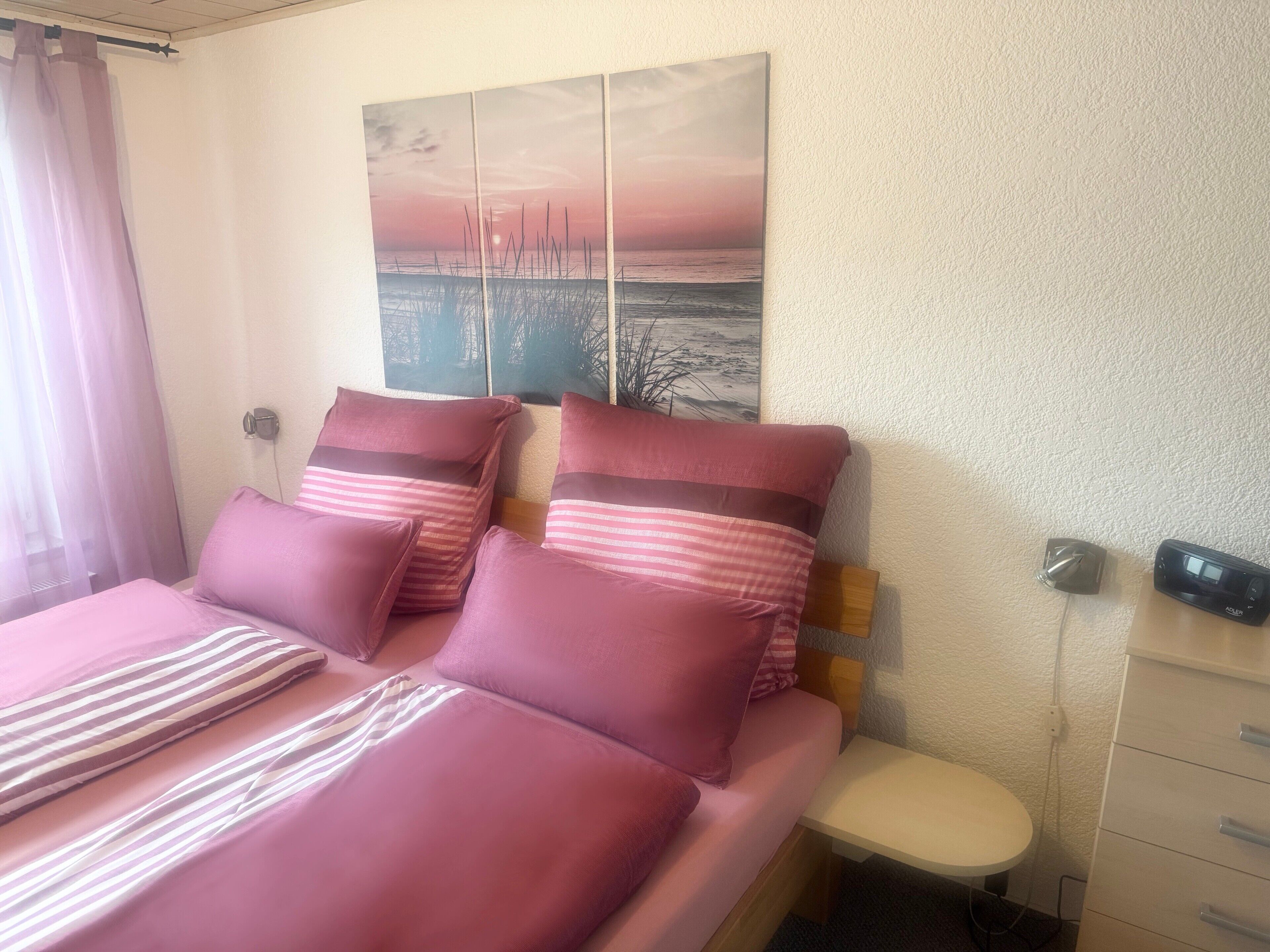 A bed with pink bedding, a bedside table, a lamp, and a wall-mounted painting.