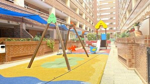 Een speelruimte voor kinderen met schommels en een glijbaan, omgeven door een gebouw met bakstenen muren en meerdere verdiepingen en balkons.