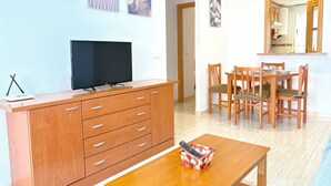 Een woonkamer met een televisie op een houten kast, een eettafel met stoelen, en een keuken zichtbaar door een open deur.