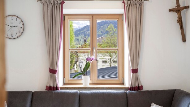 Ein Zimmer mit Holzfenster, einer Uhr, einem Kreuz und einer Couch mit Kissen.