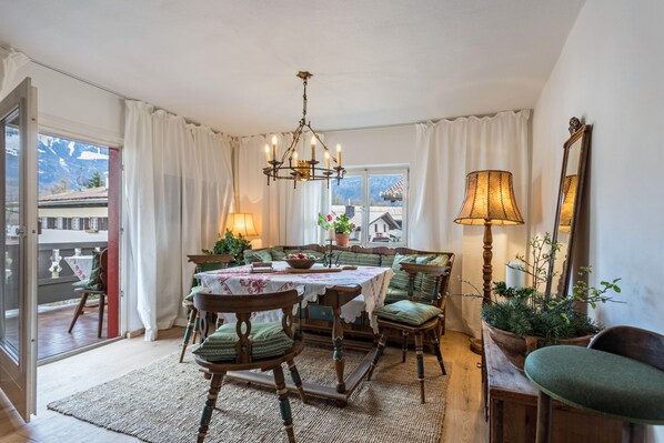 A dining room with a table set for four, surrounded by wooden chairs with green cushions, a chandelier, and a large window with curtains.