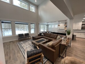 A spacious living room with a large brown sectional sofa, a coffee table, and a rug. There are multiple windows providing natural light and a view of a snowy landscape. A kitchen with white cabinets and stainless steel appliances is visible in the background. - New Yellowstone Island Park Lake House, Boat Slip, Hot Tub Sleeps 19 (Island Park)
