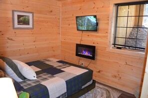 Una habitación tipo cabaña de madera con una cama, un televisor fijado a la pared y una ventana con persianas.