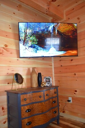 Interior de una cabaña de madera con un televisor en la pared mostrando una escena natural, un aparador de madera con cajones, un espejo y un objeto decorativo.