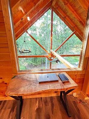 Interior em madeira com uma janela ampla, uma mesa de madeira com um laptop e um livro, e um candeeiro pendente.