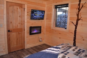 A cozy cabin room with a wooden door, a fireplace, a television displaying 'Hocking Hills, Ohio, USA', and a window with a view of snow-covered trees.
