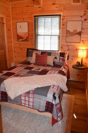 Quarto de cabana de madeira com uma cama, uma janela com persianas, uma mesinha de cabeceira com abajur e duas obras de arte na parede.