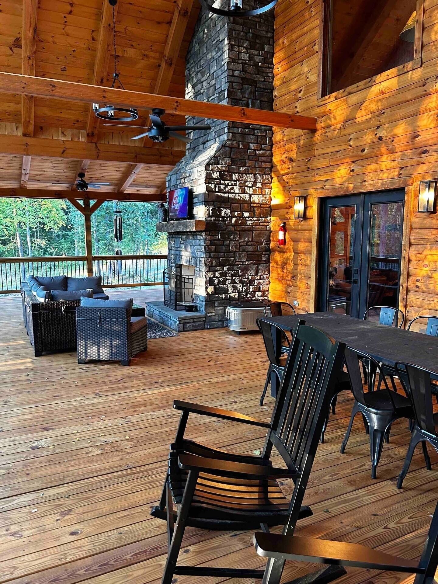 A covered outdoor deck with a stone fireplace, wooden furniture, and a view of the surrounding forest.