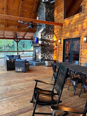 A covered outdoor deck with a stone fireplace, wooden furniture, and a view of the surrounding forest.