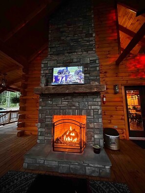 A stone fireplace with a TV mounted above it, a fire burning inside, and a wooden deck outside.