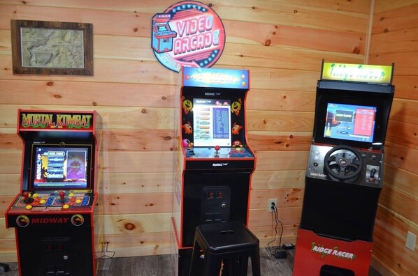 Three arcade machines labeled 'Mortal Kombat', 'Arcade Up', and 'Bridge Racer' in a wooden room.