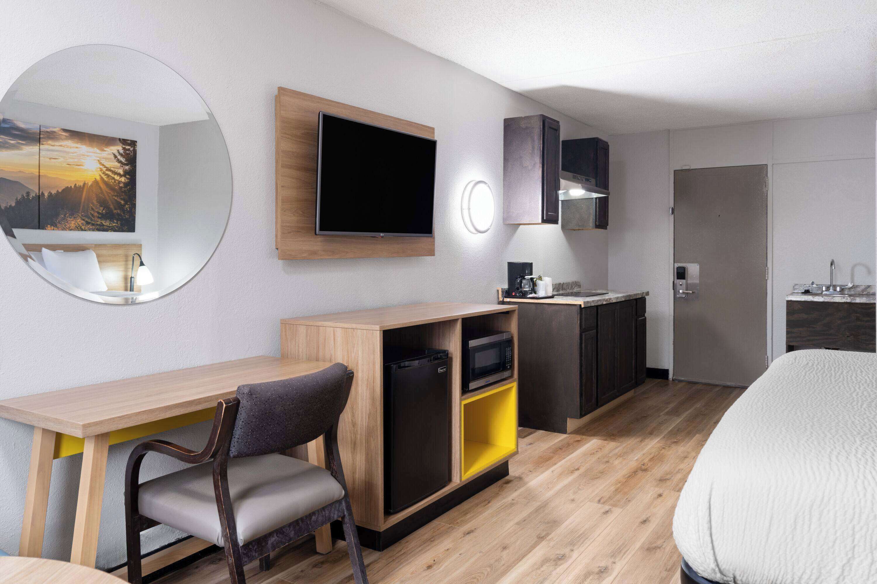 A hotel room with a wooden desk, a chair, a bed, a TV, a microwave, a mini-fridge, a sink, and a mirror.