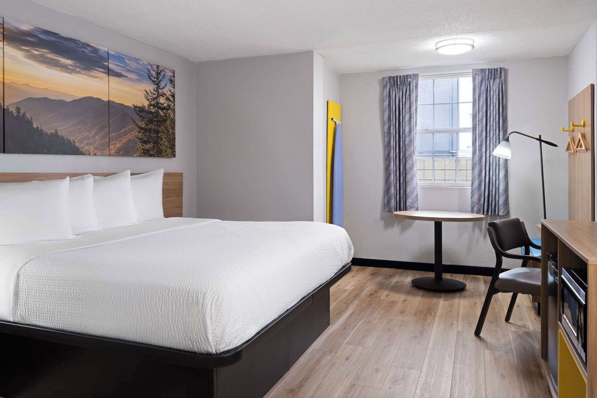 A hotel room with a bed, a desk, a chair, a table, a window with curtains, and a painting on the wall.