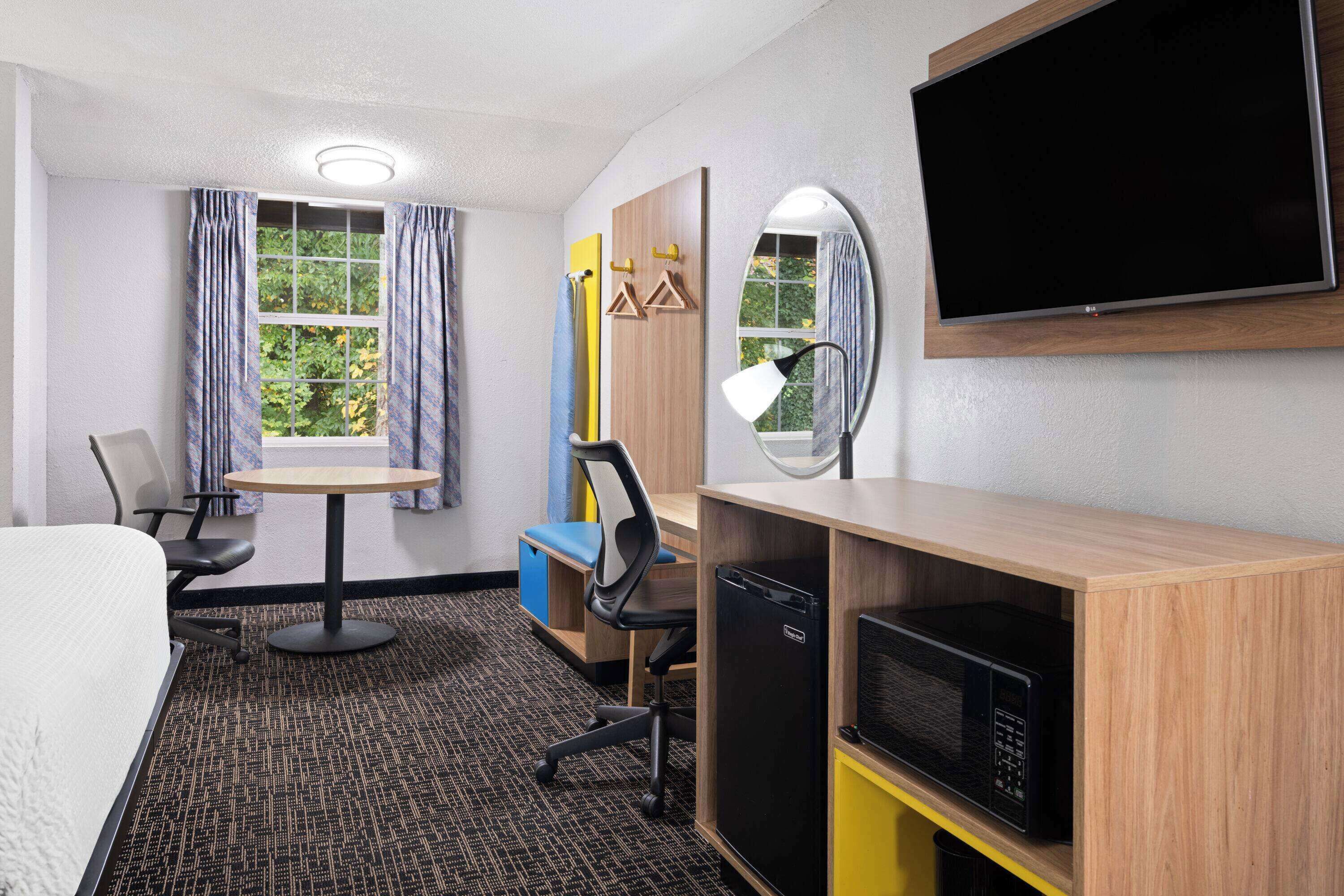 Quarto de hotel com cama, escrivaninha com computador, micro-ondas e televisão sobre uma estante de madeira.