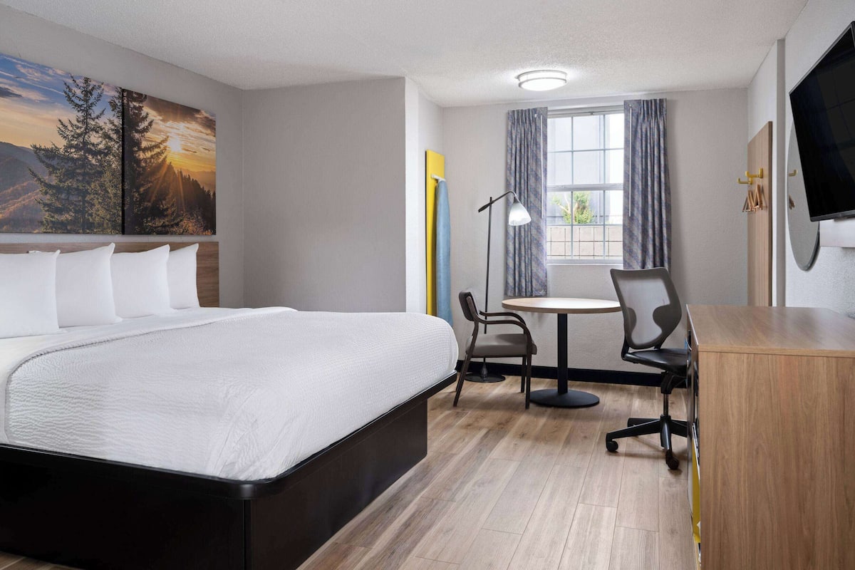A hotel room with a large bed, a desk with a chair, a flat-screen TV, and a window with curtains.
