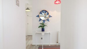 Un pasillo con una puerta blanca, una pequeña mesa blanca con planta en maceta, un tapete colorido, un espejo redondo con diseño de estrella azul y una lámpara colgante roja.