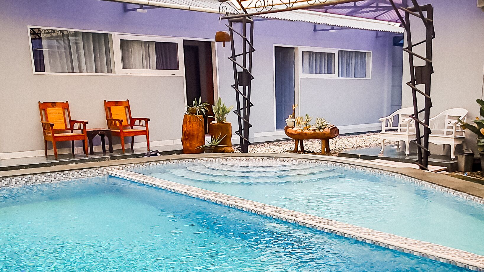 A swimming pool with a tiled edge, wooden chairs, and potted plants.