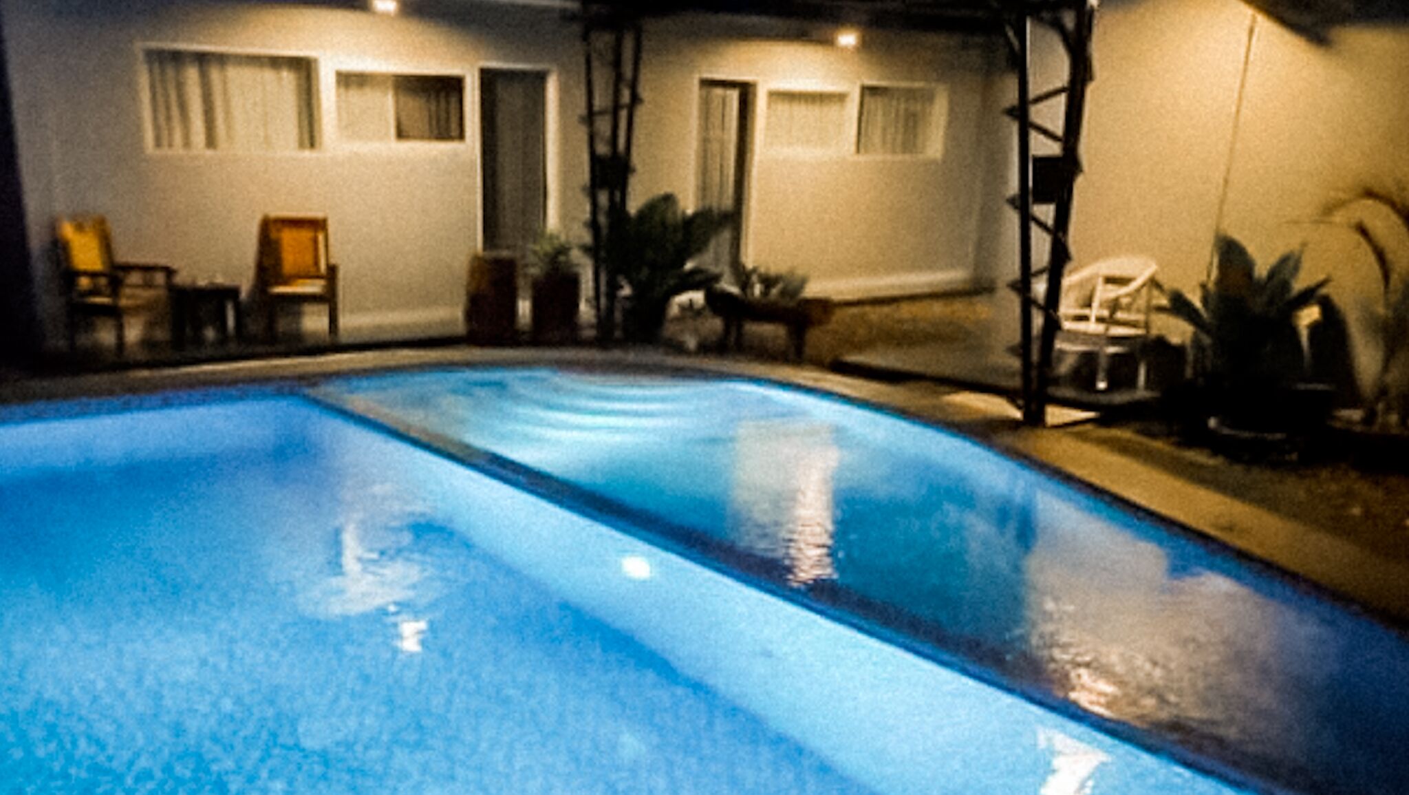 A swimming pool with a clear blue water surface, surrounded by a patio area with chairs and tables.
