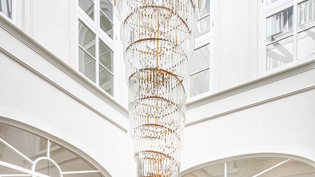 Un intérieur somptueux, dominé par un grand lustre décoratif suspendu à une colonne centrale, le tout entouré de murs blancs et de grandes fenêtres en arc de cercle.