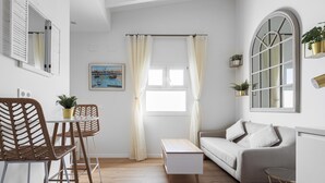 Ein modernes Wohnzimmer mit einer Couch, einem Esstisch mit Stühlen, einer kleinen weißen Kommode und einem Fenster mit Vorhängen.