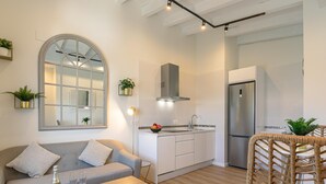 Ein modernes Wohnzimmer mit einem grauen Sofa, einem Holztisch und einem Korbstuhl. Ein Küchenbereich mit weißen Schränken und einem Edelstahlkühlschrank ist sichtbar.