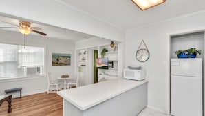 Une cuisine moderne avec des meubles blancs, un four à micro-ondes encastré et un réfrigérateur. On y trouve un coin repas avec une table et des chaises en bois, et un ventilateur de plafond au-dessus.