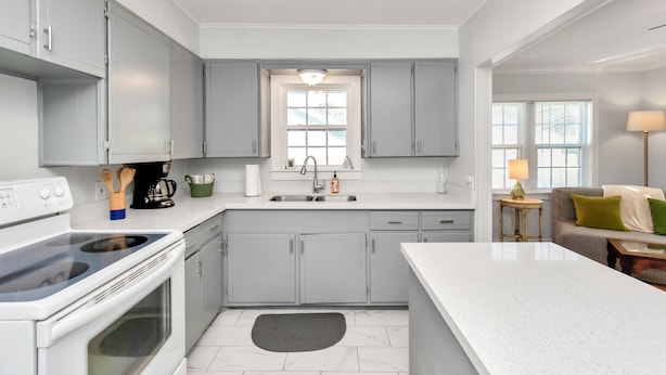Une cuisine moderne avec des meubles gris, un plan de travail blanc, un four encastré et une fenêtre au-dessus de l’évier.