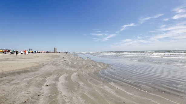 Una amplia playa arenosa con olas suaves, cielo despejado y edificios a lo lejos.
