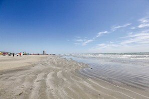 A wide sandy beach with gentle waves, a clear sky, and distant buildings. - Three Bungalows to Spread out for Large Group or Family (Galveston)
