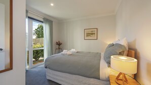 A bedroom with a bed, a bedside lamp, a window with curtains, a framed picture on the wall, and a small table with a vase of flowers.