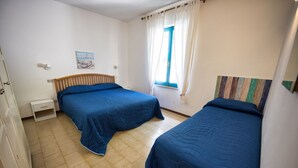 A bedroom with two beds, a small bedside table, a window with curtains, and a wall-mounted painting. - ALLOGGI DEL MIRTO - BILLY DEL MIRTO (Capoliveri)