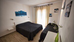 A bedroom with two beds, a wooden floor, a window with curtains, and wall art. - ALLOGGI DEL MIRTO - TRILLY DEL MIRTO (Capoliveri)