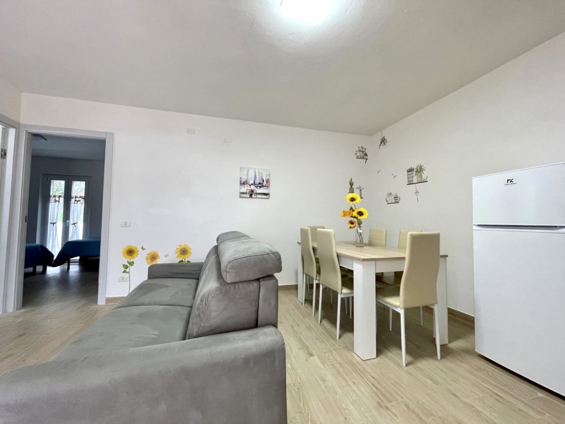 A modern living room with a grey sofa, a dining table with chairs, a white refrigerator, and a hallway leading to a bedroom.