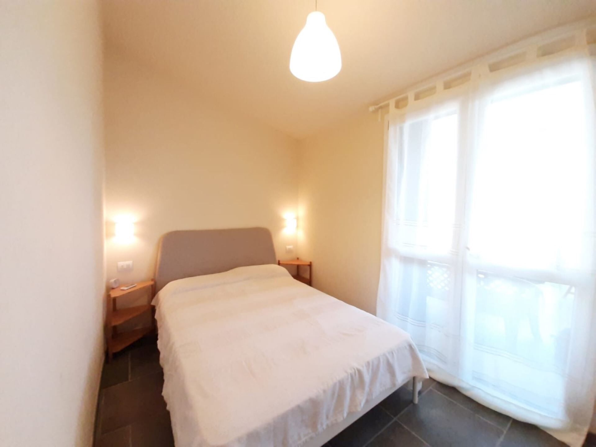 Ein kleines, aufgeräumtes Schlafzimmer mit einem Einzelbett, zwei Nachttischlampen und einem großen Fenster mit durchsichtigen Vorhängen.