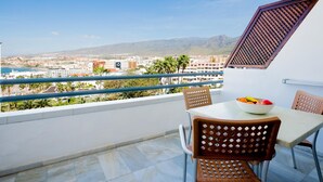 A balcony with a table and chairs, a bowl of fruit, and a view of a cityscape with mountains in the background.