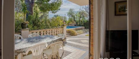 Un patio exterior con una mesa de mármol y sillas blancas, una piscina y vistas a árboles y follaje.