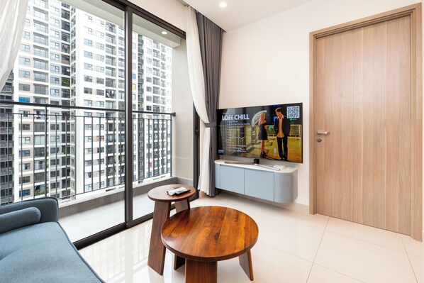 A modern living room with a sofa, a wooden coffee table, a TV mounted on the wall, and a view of a high-rise building through large windows.