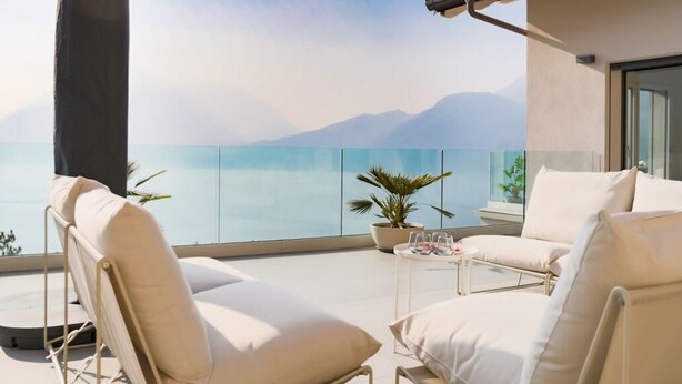 Een balkon met uitzicht op zee, twee witte fauteuils met kussens, een bijzettafeltje met glazen en een bloempot.