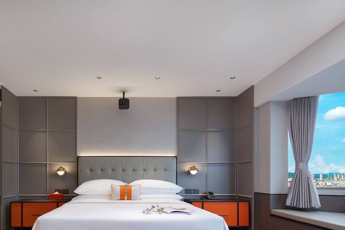 Une chambre d’hôtel moderne dotée d’un grand lit, de deux tables de chevet, d’une tête de lit avec éclairage intégré et offrant une vue sur la ville grâce à une grande fenêtre.