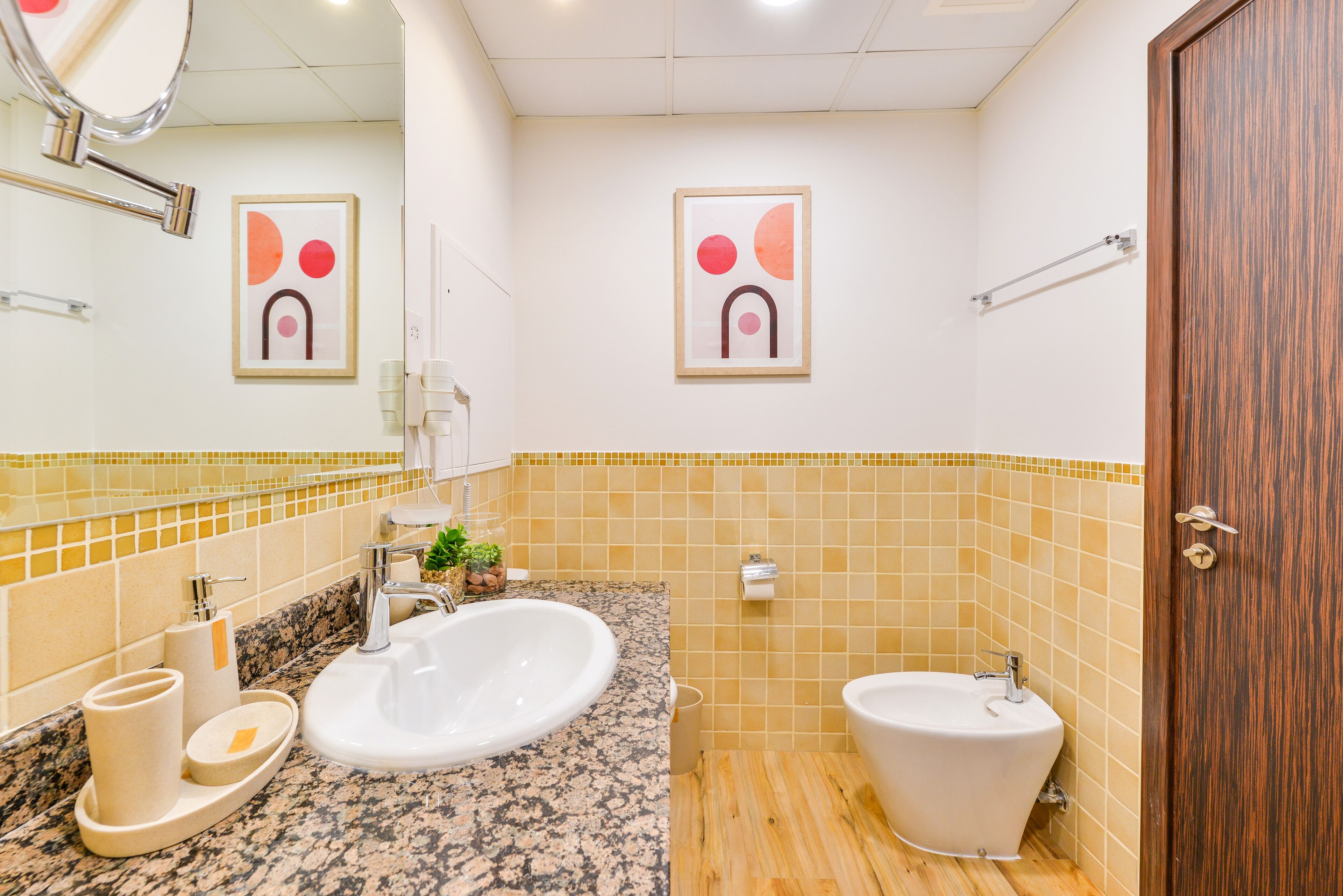 A bathroom with a granite countertop, a large sink, a bidet, and two framed abstract paintings on the wall.