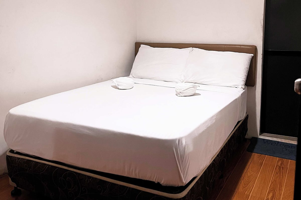 Ein Einzelbett mit weißer Bettwäsche, einem Holz-Kopfende und einem dunkel gemusterten Bettvorhang.