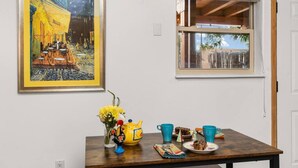 A dining area with a table set for two, featuring a vase of flowers, a teapot, and a plate of food. A painting of a café scene hangs on the wall, and a window provides a view of the outside.